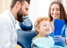 Child smiling in mirror next to dentist and dental assistant