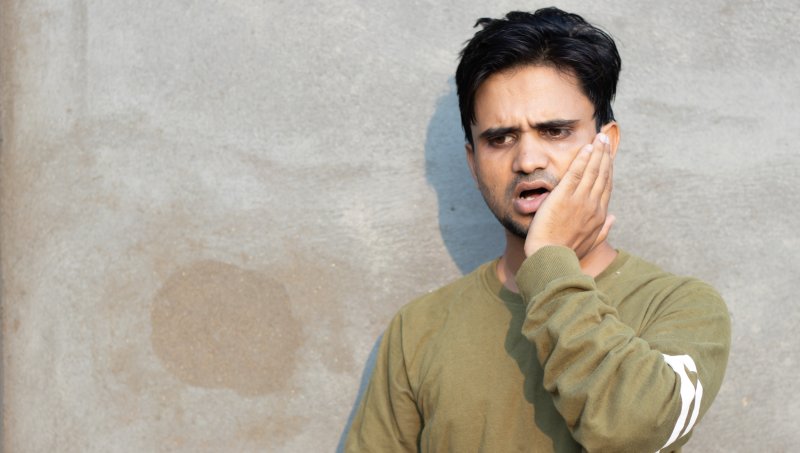 Indoor shot of young male feeling pain, holding his cheek with his hand